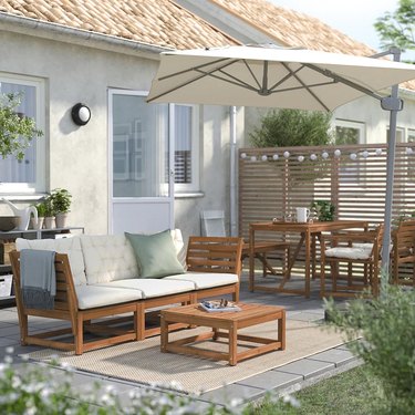 A wooden outdoor couch with pastel pillows under an umbrella in the backyard of a beige house.