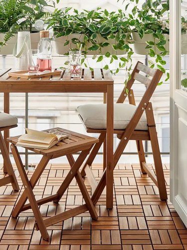 A wooden table from IKEA with wooden chairs on a patio in front of plants.