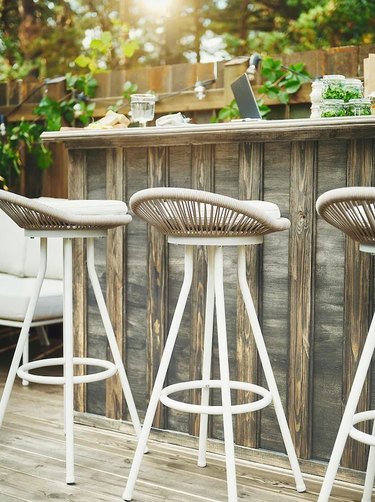 A white stool with a wicker seat in front of a wooden outdoor bar.