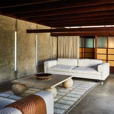 A living room with a white sofa, a travertine coffee table, a wood beam ceiling, and yellow and orange screened sliding doors.