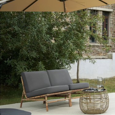 A gray outdoor sofa next to a wicker coffee table under a tan umbrella in a backyard.