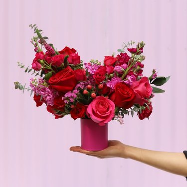 A hand holds a hot pink vase with an elaborate bouquet of red and pink roses, with greenery, berries, and smaller pink flowers throughout.