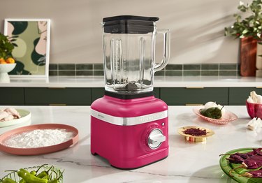 A Hibiscus KitchenAid blender rests on a kitchen counter, amidst a shallow dish of flour, a plate of raw chicken, two dishes of green and red chilis, a gold dish of red spices and a dish of onions and green peppers.