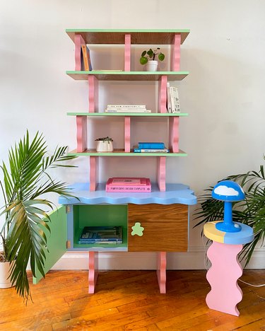 A colorful blue, green, and pink bookshelf with wavy details next to a blue, yellow, and pink side table with a wavy base that is topped with a blue IKEA cloud lamp.