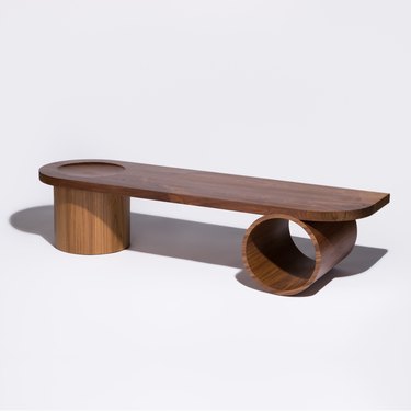 A long dark wood bench with one leg being thick and chunky, and the other being an open horizontal cylinder.