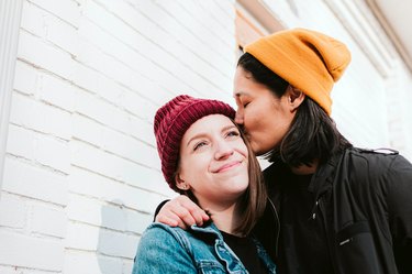 Two women wearing red and yellow beanies. One is kissing the other's forehead.
