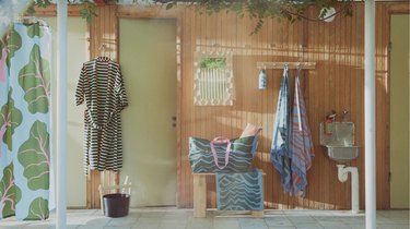 A bathroom with wooden walls, rhubarb print shower curtain, a robe hanging on a hook and a large bag on a bench