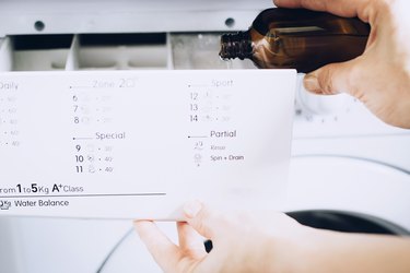 Hand pouring hydrogen peroxide from small brown bottle into third compartment of detergent compartment of white washing machine