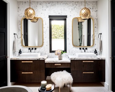 Double vanity glam bathroom with gold pendant lights, marble backsplash, sheepskin stool