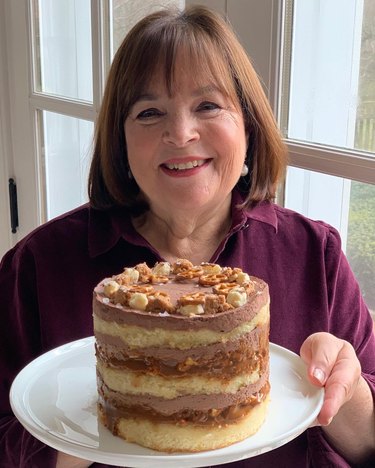 Ina Garten wearing a dark magenta collared shirt and holding a plate topped with a layered vanilla cake with chocolate frosting.