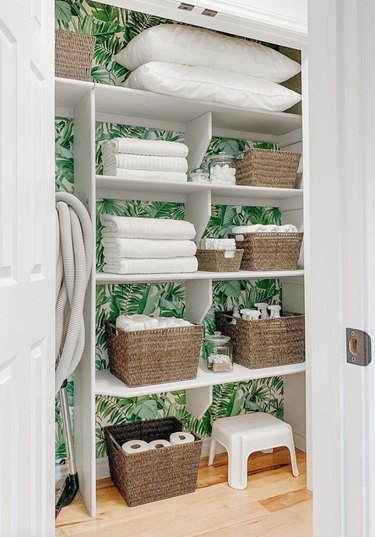 Linen closet with green wallpaper, baskets, towels, stool.
