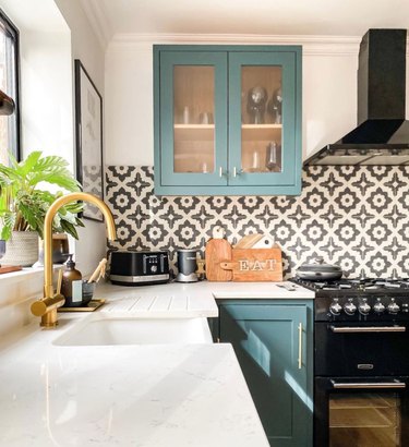 Kitchen with teal cabinets, black and white tile backsplash, black stainless steel range and hood, white counters, brass faucet.
