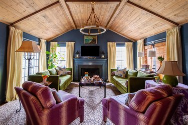 Living room with wood clad ceilings, blue walls, yellow curtains, leopard print carpet, purple chairs and green sofas.