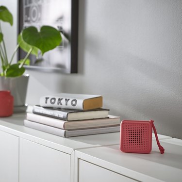 An IKEA VAPPEBY red speaker rests on a white console, next to a stack of books and a plant.