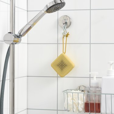 An IKEA VAPPEBY yellow speaker hangs in a white tiled shower above a shelf of shower toiletries.