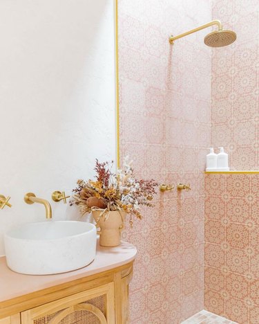 Blush, white and light wood boho bathroom with mosaic tiled shower, brass hardware, and wood and rattan vanity with terra cotta pot of dried flowers
