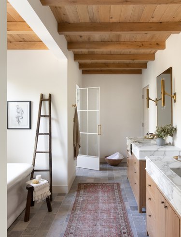 Modern rustic boho bathroom with wood beams, gray tiled flooring, white marble and wood vanities, brass accents, vintage Persian rug and wood ladder and stool