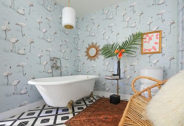 Boho eclectic bathroom with clawfoot tub, brass and white pendant light, bird wallpaper, black and white geometric tile floor, Persian rug, and woven bench with sheepskin throw pillow
