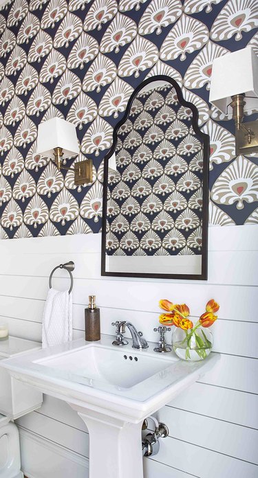 Boho bathroom with white shiplap along the bottom of the wall and an Indian print wallpaper along the top half, Moroccan vanity mirror with double sconces and simple white sink.