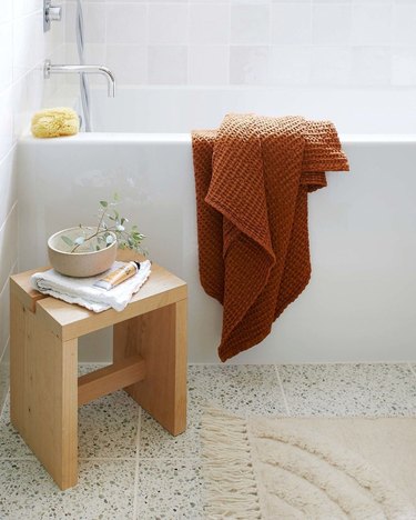 warm neutral boho bathroom with caramel towel thrown over a white bathtub, small light wood stool, and cream colored fringe rug