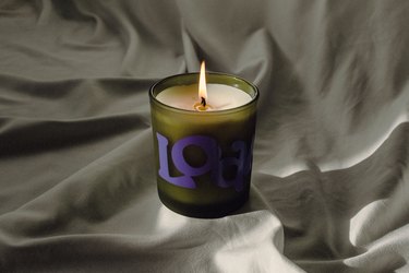 A lit Loam candle in a green jar with purple logo on a wrinkled sheet.