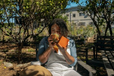 Jessica White smelling one of her candles and holding its orange box on a wood chair in the midst of a community garden.