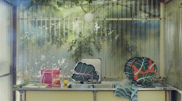 Rhubarb leaf-themed plates, towels, and a pink tote bag on a metal countertop in front of a semi-transparent wall with outdoor greenery behind it.