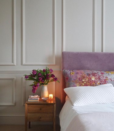 Bedroom with lilac upholstered headboard, board and batten walls, nightstand, flowers, bedding.