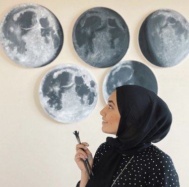 Painter Zarah Khan stands in front of five circular moon paintings and is holding a paintbrush