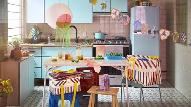 A colorful kitchen featuring IKEA's ÖMSESIDIG collection. A paper lantern and garland are hung from the ceiling, the kitchen tables has assorted glasses of juice and limes. Colorful bags rest on the dining chairs, and a colorful card sits atop a wooden stool.