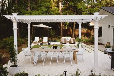 A large white pergola on white gravel. There is a large wooden dining table underneath, with room for six white chairs. An additional seating area is behind pergola.