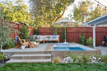 A backyard patio with lush plantings and a small plunge pool. A white and blue inner tube floats on the water, and there is a white umbrella and outdoor seating nearby.