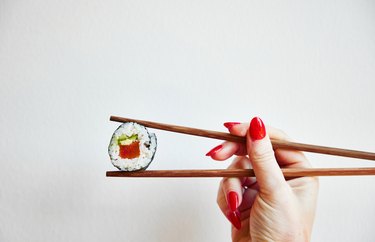 A person with red nails holding chopsticks with plant-based sushi in the center of the chopsticks.
