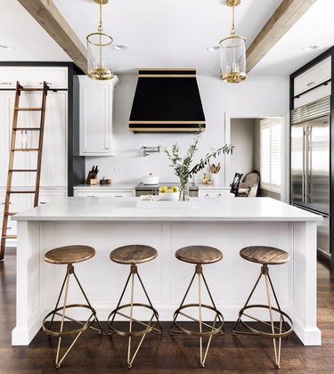 Kitchen with white island, marble counter, brass bar stools, black and gold hood, glass pendants.