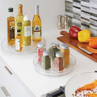 Two plastic turntables rest on a white kitchen counter, one filled with cooking oils, and the other filled with spices.