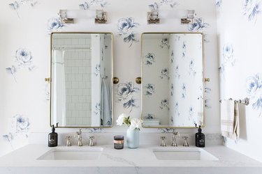 white bathroom with silver fixtures, gold mirrors, and white and blue floral wallpaper