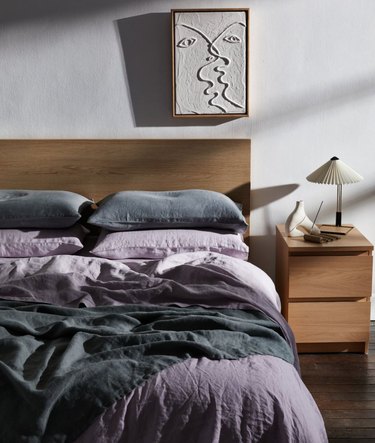 Bedroom with wood headboard, art, lilac and gray bedding, nightstand, lamp.