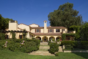 Cream mission-style home red-tile roof, sprawling green lawn, and paved stone walkway