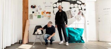 A white dog on a black chair is being petted by a man with short gray hair crouched down on the floor next to a man with short brown hair and beard wearing circular glasses in front of a white mood wall of fabric samples and designs.