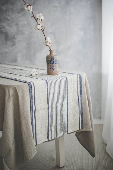 A blue and cream striped tablecloth on a table topped with a vase and flowering branch.