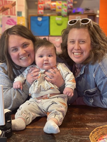 Two women with medium-length brown hair are smiling and holding an adorable baby with short brown hair sitting on a dark wood table. In the background are colorful storage boxes and sticker rolls on display.