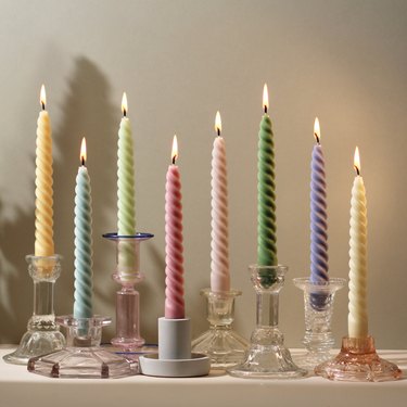 Twisted candles in various colors, lit and places in various candle holders on a white surface.