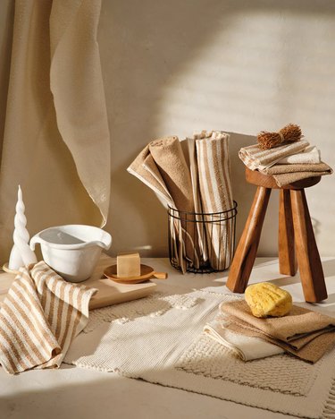 Beige and white striped and solid towels are folded and rolled in a bathroom setting with wood stool and tray, black wire basket, and white ceramic pitcher