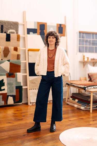 Woman with curly, shoulder-length brown hair standing with hands in pockets wearing a white cardigan sweater, rust shirt, blue pants and black boots in a room with hardwood floors and hanging patterned fabrics in the background