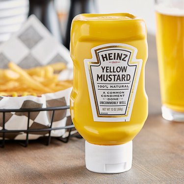 A container of Heinz yellow mustard on a wood table in front of a basket of fries.
