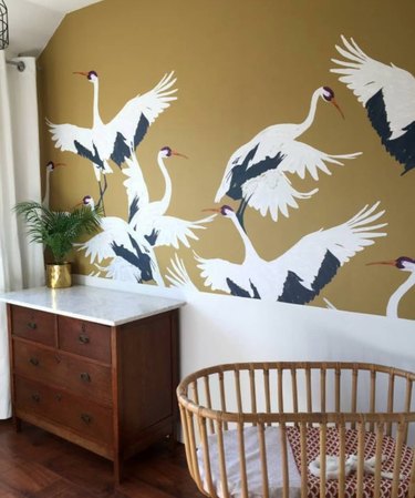 Nursery with cane crib, wood dresser, wallpaper with stork pattern.
