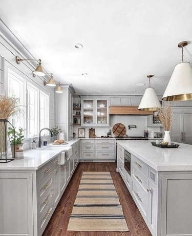 A bright and open kitchen, with light grey cabinets, gold and white lighting fixtures, gold cabinet pulls, a matte black faucet, glass cabinet doors, a wood range hood over the oven and a jute runner with navy blue stripes on the floor.