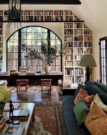 Traditional living room with home library, books, couch, pillows, coffee table, dining table, chairs, rug.