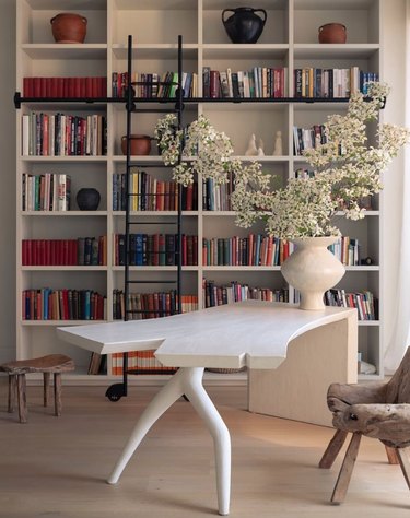 Modern living room with home library, books, table, chair, vase, ladder.