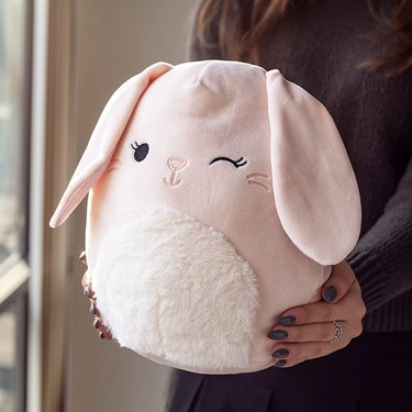 A round light pink plush bunny toy with a white fuzzy stomach being held by someone with gray painted nails.